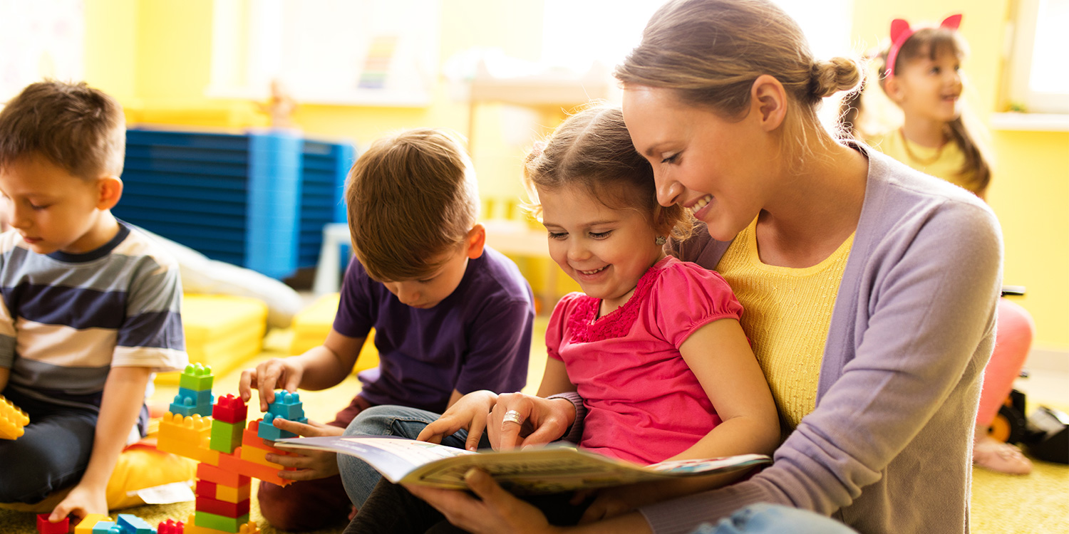 Kinderkrippenalltag. Kinder spielen und eine Pädagogin liest aus einem Buch vor.